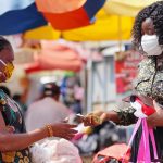 Young black woman paying money to a vendor for a purchased item in the market. Two women wearing locally made mask and surgical mask on the street.